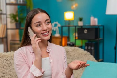 Kanepede oturan genç bir kadın akıllı telefonda konuşurken gülümseyip el sallıyor ve arkadaşça sohbet ediyor. Kafkasyalı kız mutlu sıcak tavırlar sergiliyor. Gülüyor ve uzak bir arkadaşla pozitif enerjiyi paylaşıyor..