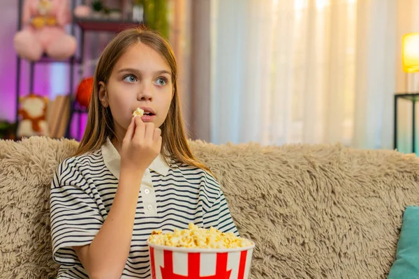 Genç kız patlamış mısır yer ve kanepede oturup eğlenirken televizyonda komik çizgi filmler izler. Okullu çocuk evde rahat bir film izlerken keyifli ve rahat görünüyor.