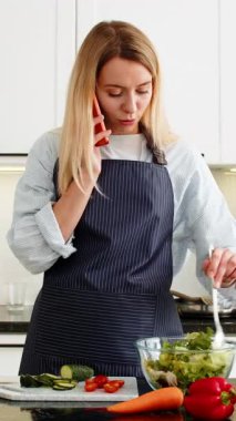 Orta yaşlı bir kadın, mutfakta sebze salatasını karıştırırken turuncu akıllı telefonla konuşuyor ve vejetaryen tarifini paylaşıyor. Genç kız sağlıklı ev yemeği öğreniyor. Kilo kaybı ve beslenme alışkanlığı için hazırlık yapıyor.