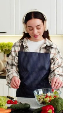Genç bir kadın salata kasesine domates ve kulaklıkla ev mutfağında sakin müzik dinliyor. Kız, evde temiz beslenmek için taze sebzelerle vejetaryen yemeği hazırlıyor.