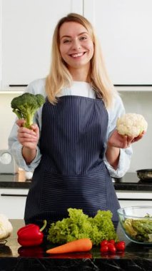 Orta yaşlı bir kadın mutfakta brokoli ve karnabahar tutuyor. Keto diyeti ve kilo vermek için sağlıklı bakkal taşımacılığı yapıyor. Genç kız düşük karbonhidratlı yemek hazırlamayı evde video blogundan öğreniyor ve gülümsüyor.