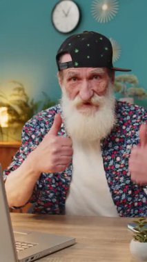 Gibi. Gülümseyen stil sahibi yaşlı adam kameraya olumlu bakıyor. İki başparmak işareti gösteriyor. İyi haber olumlu geri bildirim. Ev ofis masasında dizüstü bilgisayarı olan mutlu büyükbaba..