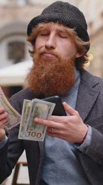 Mutlu genç adam dolar sayıyor, akıllı telefon hesap makinesi uygulaması kullanıyor, planlanmış tatil hediyeleri için gelirden tatmin oluyor. Şehir sokağındaki kafede oturan kızıl saçlı çocuk. Şehir yaşam tarzları açık havada