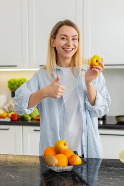 Orta yaşlı bir kadın mutfak masasında taze elma yiyor. Vitamin ve diyet için sağlıklı atıştırmalıkların tadını çıkarıyor. Genç kız, günlük temiz yemek ve sağlık rutinini destekleyen tatlılar yerine meyve seçiyor..