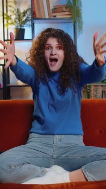 Kıvırcık saçlı mutlu genç bir kadının portresi başparmak dansı göstererek başarıyı kutluyor evdeki oturma odasındaki kanepede oturuyor. Evde maç izlerken rahat giyinen heyecanlı bir yetişkin kız..