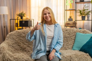 Jest gibi. Genç bir kadın, başparmağını kaldırmış, dostça bir anlaşma ve destekleyici bir kafa sallama ile evde oturuyor. Kanepedeki kız günbatımının parlaklığı altında basit bir memnuniyeti doğrulayan sıradan bir gülümseme sergiliyor.