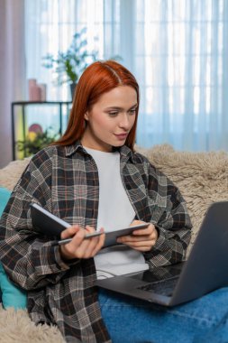 Evde genç bir kadın odaklanarak, başını sallayarak ve not defterine notlar yazarak çevrimiçi derse katılıyor. Kanepede oturan beyaz saçlı kız aktif olarak öğreniyor, eğitime ve sanal sınıfa odaklanıyor.