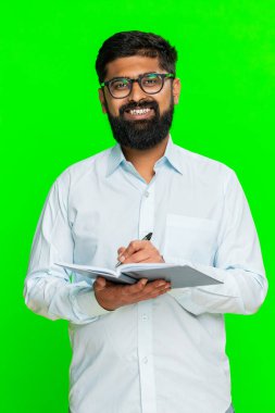 Hintli adam defterine ciddi odaklanmış bir ifadeyle günlük işleri planlar ve yapılacaklar listesi düzenler. Arap adam yeşil krom anahtar arka planda izole edilmiş. Konsantrasyon ve hazırlık gösteriyor.