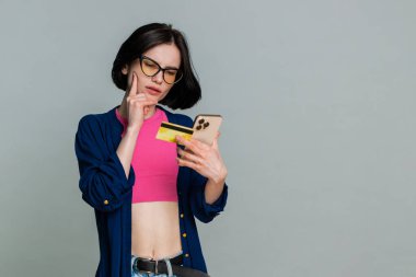 Kredi kartı ve akıllı telefon kullanan mutlu bir kadın. Harcama parasını transfer ederken, online alışveriş yaparken, yemek siparişi verirken. Kız, gri arka planda izole edilmiş satın alma konusunda kendinden emin görünüyor