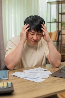 Masada oturan Asyalı adam kafasını tutuyor, ödenmemiş faturalar hesap makinesi ve dizüstü bilgisayarın yanına saçılmış. Çinli adam derin bir iç çekiyor, ağır mali stres gösteriyor ve ev bütçesini acilen dengeleme ihtiyacı duyuyor..