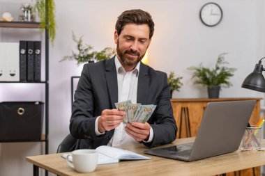 Merkez ofisteki orta yaşlı işadamı nakit para sayıyor ve aylık bütçesi için dizüstü bilgisayardan giriş yapıyor. Masadaki serbest çalışan adam not defterine notlar yazıyor. Hesaplar, harcamalar ve tasarruflar ekliyor..