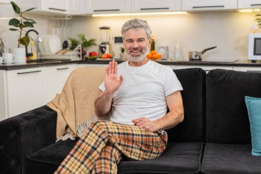 Evde kameraya el sallayan orta yaşlı bir adam merhaba diyor ve arkadaş canlısı gülümsemelerle çevrimiçi iletişim kurmaya başlıyor. Kanepedeki olgun adam lenslere bakar kısa selam verir sıcak hava konuşur açık ton sakin nefes al istikrarlı.