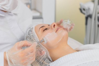 Enzim terapisi. Güzellik uzmanı orta yaşlı bir kadının yüzüne fırçayla nemlendirici kimyasal krem maske takıyor. Yetişkin kız cilt bakımı prosedürü. Yüz kozmetolojisi tedavisi.