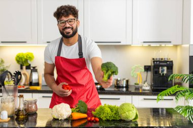 Arap adam, brokoli ve kırmızı biberi mutfakta diyet beslenmesi için sağlıklı yemek hazırlarken karşılaştırıyor. Genç adam gülümseyerek ev yemekleri ve fitness yaşam tarzı için taze sebze seçiyor..