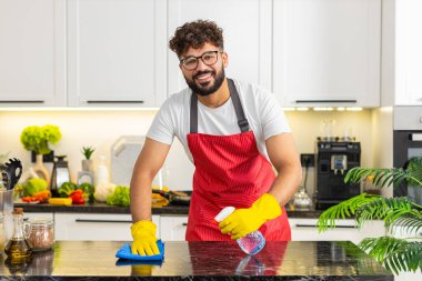 Arap adam mutfak tezgahını mikrofiber bezle temizliyor. Temiz ev hijyeni ve gıda güvenliği için leke çıkartıyor. Ev işi yapan genç adam, ev mutfağını düzenli tutmaktan memnun ve odaklanmış hissediyor..