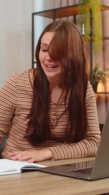 Evde genç bir kadın kulaklıkla video görüşmelerine katılıyor elleriyle el kol hareketleri yapıyor ve not defterine notlar yazıyor. Masadaki beyaz kız dizüstü bilgisayara bakıyor. Dinliyor, dakikalar alıyor ve konuşma sırasında odaklanıyor.