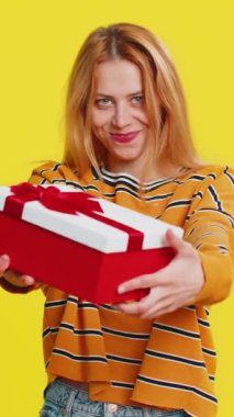 Doğum günü hediyelerini sunan mutlu ve mutlu genç bir kadın ellerini uzatıyor, hediye paketine sarılmış kariyer ikramiyesi teklif ediyor, parti promosyon indirimi Noel indirimini kutluyor. Sarı arka plandaki kız