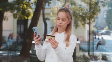 Para transfer ederken kredi kartı akıllı telefonu kullanan gülümseyen bir kadın, online alışveriş, yemek siparişi, açık havada otel rezervasyonu. Şehir caddesinde duran genç kız..