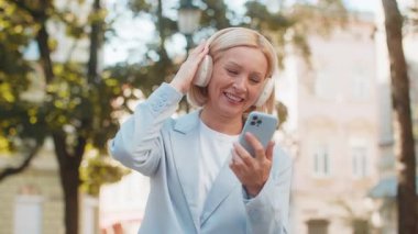 Neşeli, kaygısız, olgun, beyaz iş kadını şehir sokaklarında kulaklıkla müzik dinliyor. Mutlu orta yaşlı kadın elinde akıllı telefon dansı tutarken şarkı dinleyerek şehirde dolaşıyor..
