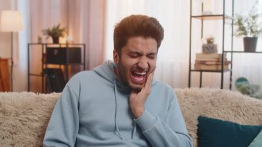 Diş sorunları. Yanağa dokunan genç Hintli adam, korkunç bir ifadeyle gözlerini kapatıyor diş ağrısı, hassas dişler, çürükler yüzünden acı çekiyor. Hintli adam evdeki kanepede oturuyor..