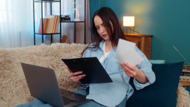 Evdeki mutlu genç kadın, mali faturaları ve bilgisayardaki belgeleri incelerken gülümsüyor ve iyi bir jest yapıyor. Kanepedeki beyaz kız ekonomik sonuçlardan memnun, olumlu bütçe duyguları var.