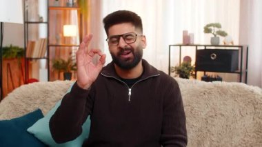 Evdeki Hintli adam kanepeye oturur ve rahatça gülümser, kameraya hoş bir jest yapar. Arap adam iyimserliğini ve pozitif ruh halini ifade ediyor, kendine güveni yansıtıyor ve kapalı alanda rahat bir enerji veriyor..