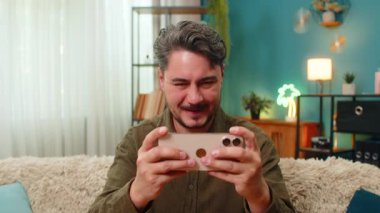 Evde orta yaşlı bir adam akıllı telefon oyununu yener piyango vurur kollarını havaya kaldırır ve büyük sevinçli zafer diye bağırır. Kanepede oturan genç adam cep telefonuyla dans ediyor. Heyecanın tadını çıkarıyor. Gurur kazandı.