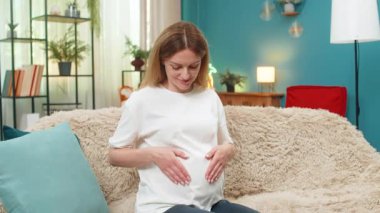 Hamile, orta yaşlı bir kadın kalbinin şeklini değiştiriyor. Elleri karnında ve gülümsemeleri saf sevgi sıcaklığıyla dolu. Kanepede oturan beyaz kız bebekle duygusal bir bağ hissediyor. Sakin, tatlı, büyük bir sevinç anı yaşıyor..