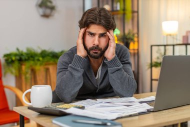 Evdeki Hintli adam el ele tutuşup hesap makinesi ve dizüstü bilgisayarla ödenmemiş faturaları görüyor. Masa başı kontrolleri yapan Arap adam tarih sayımı maliyetli ödeme düşük bütçe endişeli ve sakin nefes.