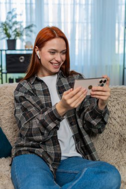 Genç bir kadın akıllı telefondan video izleyip kulaklık takıyor ve güçlü duygularla gülüyor. Kafkasyalı kızıl saçlı kız kanepenin üzerinde eğleniyor, neşe ve neşeyle tepki veriyor.
