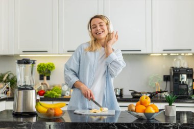 Orta yaşlı bir kadın beslenme için mutfakta kulaklıkla dans ederken blender 'da smoothie için muz dilimliyor. Genç kız müzikten hoşlanıyor ve enerji ve sağlık için taze narenciye içeceği hazırlıyor..