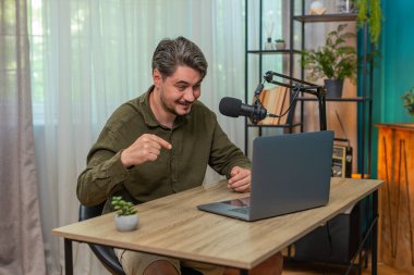 Evdeki orta yaşlı adam canlı video bloğu sunuyor. Laptop mikrofonuna sesleniyor. Seyirci etkileşimini destekliyor. Masadaki genç adam canlı konuşmalar yapıyor. İpuçlarını ekranda tutkuyla paylaşıyor.