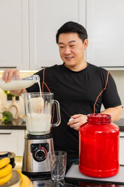 Asyalı bir adam, temiz diyet yapmak için muzlu süzme peynirli whey proteinle karıştırıcıya süt döker. Yüksek protein içeceği ev sağlığı yaşam tarzı için kas düzeltme ağırlığı kontrol enerjisini destekler.