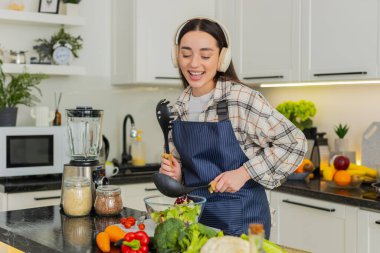 Genç bir kadın evde vejetaryen salatası hazırlarken kulaklıkla müzik eşliğinde dans ediyor ve kaşıkla kepçe takıyor. Kız gülümser ve sağlıklı vejetaryen yemeği için taze sebzeleri kaseye koyar..