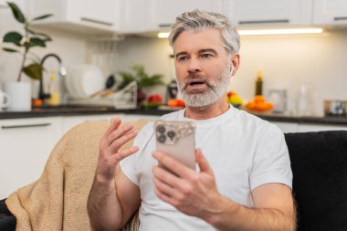 Evde orta yaşlı bir adam akıllı telefon uygulamasından kelimeleri kulaklıkla öğreniyor ve geri bildirim duyuyor. Olgun adam koltukta durur ve gülümser. Yüksek skoru görür. Büyük ilerleme kaydeder ve çalışmaya devam eder..