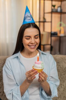 Evde elinde mumlarla mutlu bir şekilde gülümseyerek doğum günü pastası tutan genç bir kadın. Kanepedeki beyaz kız heyecanlı, duygusal olarak olumlu, neşeli bir ruh hali içinde, neşeli bir doğum günü kutlaması.