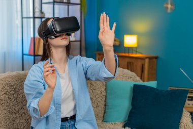 Evde VR kulaklığı takmış, kanepede oturan genç bir kadın inanılmaz bir şekilde başını çeviriyor ve hareketler yapıyor. Beyaz kız heyecan verici bir şekilde sanal gerçekliği keşfediyor, şaşkınlık ve neşe duyguları yaşıyor.