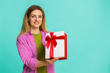 Orta yaşlı bir kadın ilginç bir şekilde bir hediye kutusunu açıyor ve içinde ne olduğuna mutlu bir şekilde tepki veriyor, heyecan, neşe ve şaşkınlık gösteriyor. Mavi arka planda izole edilmiş bir kız neşeli duygular ve hareketlerle kutluyor..