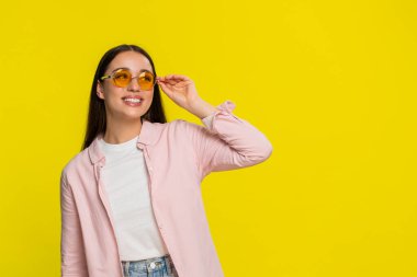 Şık bir güneş gözlüğü deneyen genç bir kadın neşeli bir ruh hali sergileyen komik bir ifadeyle neşe içinde poz veriyor. Sarı arka planda izole edilmiş bir kız neşeli bir enerji gösterisi sergiliyor.