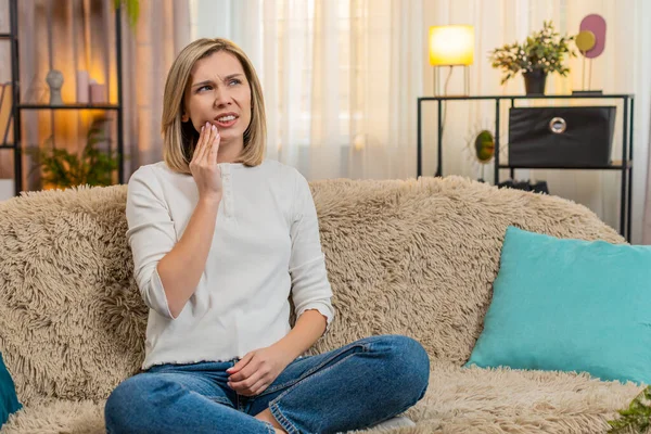 Orta yaşlı bir kadın, diş ağrısından ürkmüş bir vaziyette elinde yanağıyla kanepesinde oturuyor. Yetişkin hasta kadın gergin bir yüz ve vücut diline sahip keskin bir ağrı hissediyor. Rahatlamak ve sakinleşmek istiyor..