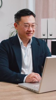 Asyalı işadamı, modern ofis çalışma masasında zafer kutlamaları yaptıktan sonra sandalyeye yaslanıyor. Resmi takım elbiseli erkek Çinli ellerini kafanın arkasına koyup rahatlatıyor.