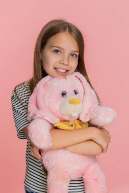 Genç kız pelüş oyuncak tavşancığa sevgi ve mutlulukla sarılıyor, sevinçle tatlı tatlı gülümsüyor, sıcaklığını ve duygusal bağını ifade ediyor. Pembe arka planda izole edilmiş küçük okul çocuğu..