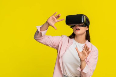 VR kulaklık takan genç bir kadın ellerini havaya kaldırıyor ve sanal dünyaya hayran kalıyor. Sarı arka planda izole edilmiş bir kız dijital deneyime olan hayranlığını ve heyecanını ifade ediyor.
