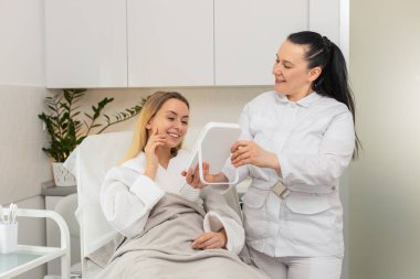 Güzellik kliniğindeki kozmetik ameliyatından sonra beyaz kadın aynada gülümsüyor. Yetişkin bir bayan görünür sonuçtan zevk alır, kendini yenilenmiş, kendinden emin hisseder, cilt bakımındaki ilerlemeden tatmin olur, profesyonel ellere güvenir..