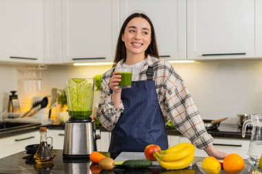 Kadın, blender kavanozundan yeşil smoothie 'yi mutfaktaki cama dolduruyor, içmeye hazır. Temiz yemek ve taze meyve ve sebzeli sağlıklı yaşam tarzı konsepti. Ev yapımı kahvaltılık içecekten hoşlanan kız..