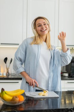 Orta yaşlı bir kadın beslenme için mutfakta kulaklıkla dans ederken blender 'da smoothie için muz dilimliyor. Genç kız müzikten hoşlanıyor ve enerji ve sağlık için taze narenciye içeceği hazırlıyor..