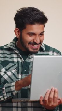 Heyecanlı mutlu Hintli genç adam dizüstü bilgisayarda yazıyor, proje üzerinde çalışıyor, sonuçların tadını çıkarıyor, piyango kazanıyor, online alışveriş başarısını kutluyor, kazanıyor. Hintli çocuk bej renkli stüdyo arka planında izole edilmiş.