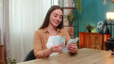 Evde oturan neşeli genç kadın UAH Ukrayna Hrivnia 'lı para hesaplarını sayıyor. Maaş kazancından memnun kalmış şekilde gülümsüyor. Kız kendini zengin hissediyor. Finansal başarıyı kutluyor, alışverişleri planlıyor.