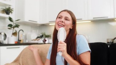 Evde genç bir kadın pop idolü fantezisi icra ediyor mikrofon olarak tarakta şarkı söylüyor ve eğlence için dans ediyor. Kanepede oturan beyaz kız gülüyor, el çırpıyor, müzik çalıyor ve neşeli bir ruh hali içinde..