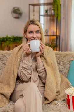 Genç, yetişkin bir kadın sıcak bir battaniyeye sarılmış, sıcak bir çay tutuyor ve sakin bir şekilde diziyi seyrediyor. Sarışın kız düşünceli görünüyor, sıcak içki ve konforla huzurlu bir anın tadını çıkarıyor..